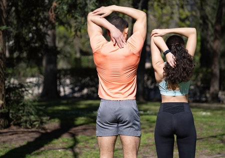 Rear view of man and woman athletes stretching arms and back in park before exercising