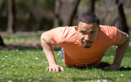Young sportsman performing push-ups on grass in a park, demonstrating strength and dedication to...