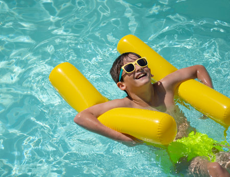 Cheerful young boy wearing sunglasses enjoys summer holidays, relaxing on yellow inflatable in swimming poolの写真素材