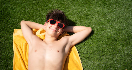 Top view of a happy young teenager boy is lying on a yellow towel in grass wearing red sunglasses and relaxing with copy spaceの写真素材