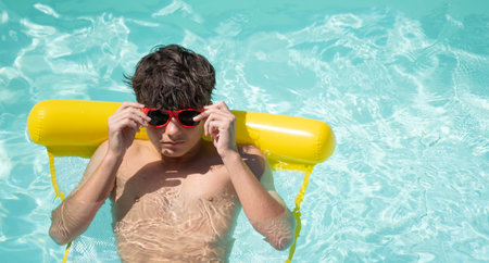 Young man enjoying summer vacation, floating on yellow inflatable mattress in turquoise pool, adjusting red sunglasses, with copy spaceの写真素材