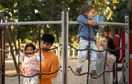 Latin parents helping children climbing on playground equipment, enjoying a sunny day at parkの写真素材