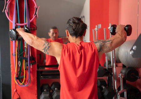 Strong man wearing red sleeveless shirt performing lateral raise exercise with dumbbells in a gymの写真素材