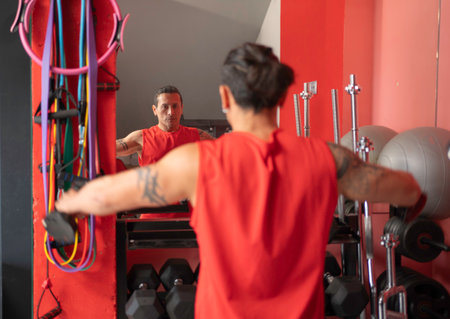 Rear view of muscular man working out with dumbbells in a gym, looking in the mirrorの写真素材