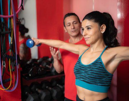 Side view of smiling latin woman exercising with dumbbells and personal trainer assisting herの写真素材