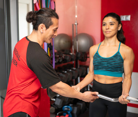 Hispanic personal trainer assisting a sporty woman during her weightlifting workout in a modern gym. Fitness and healthy lifestyle conceptの写真素材