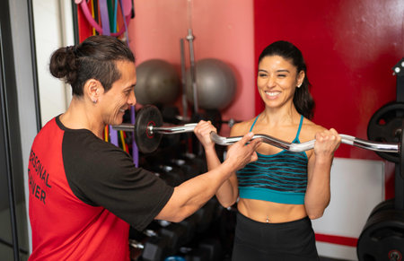 Smiling personal trainer assists a fit woman lifting a barbell in a modern gymの写真素材