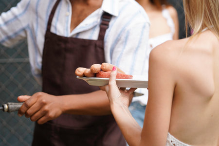 Close up of woman holding plate of grilled sausages and burgers at barbecue party, male chef is grilling in backgroundの写真素材