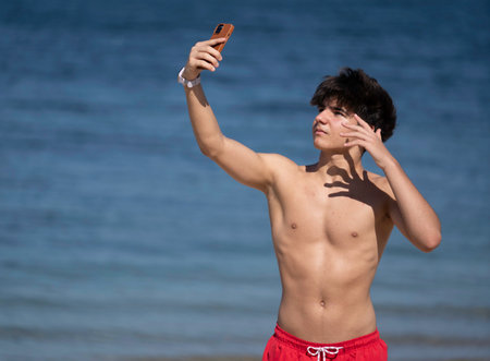 Portrait of confident young man taking a selfie with smartphone while enjoying a sunny day at the beachの写真素材