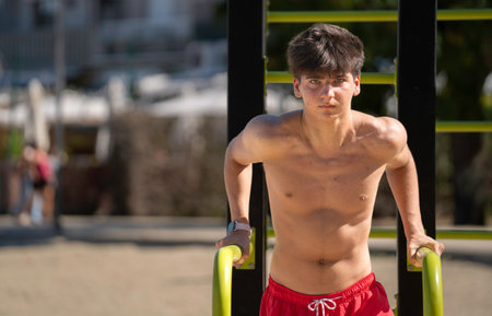 Portrait of a teenager boy exercising outdoors, strengthening his upper body using parallel bars on...