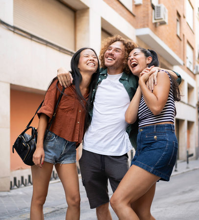 Three cheerful multiethnic young friends are embracing and laughing together on a city street,...