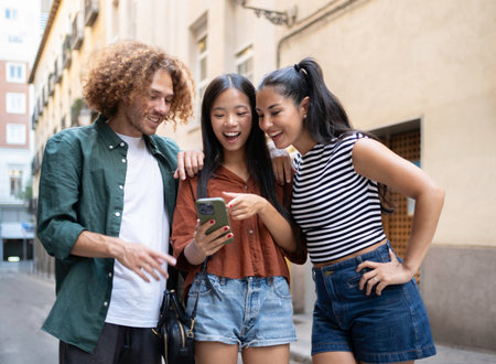 Three happy young tourists are using a smartphone and laughing while walking down a city streetの写真素材