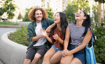 Three cheerful diverse students are sitting on a low wall, laughing and enjoying each other's...