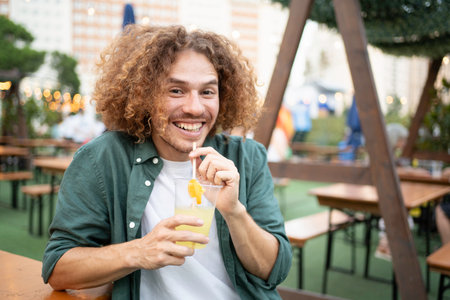 Portrait of young hispanic man with curly hair enjoys refreshing lemonade at an outdoor cafe, smilingの写真素材