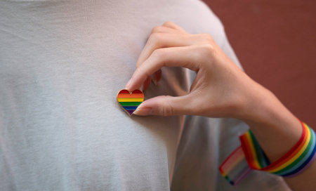 Close up of woman hand placing a rainbow heart pin on a gray t-shirt, symbolizing support for lgbtq...
