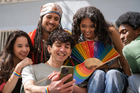 Multiethnic group of cheerful multi-ethnic young people showing lgbtq pride flag and rainbow fan, using smartphone outdoorsの写真素材