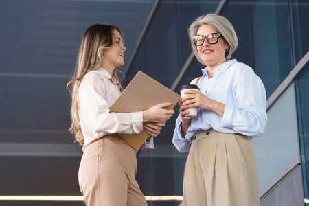 Two businesswomen are having a conversation outside their office building, holding coffee and documentsの写真素材