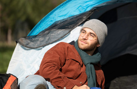 Smiling young man relaxing outside his tent, enjoying a hot beverage and contemplating the landscape