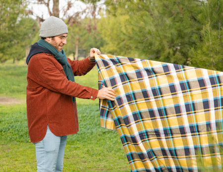 Man arranging checkered picnic blanket on meadow in park during autumn dayの写真素材