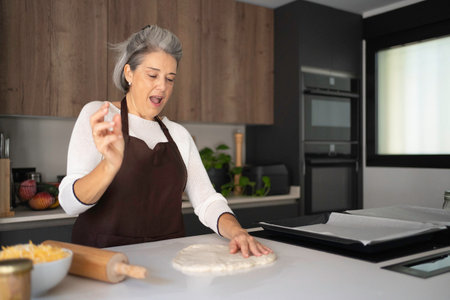 Mature woman happily preparing pizza dough on a kitchen counter, enjoying the cooking processの写真素材