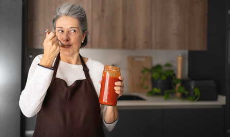 Senior woman tasting sauce from a jar, preparing food in a modern kitchenの写真素材