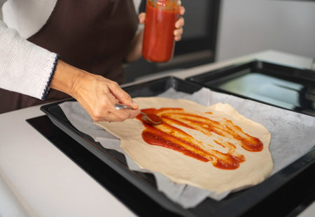 Woman's hands spreading tomato sauce from a jar onto raw pizza dough on a baking sheetの写真素材