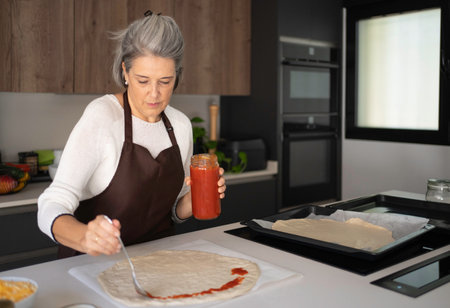 Woman spreading tomato sauce on pizza dough for a homemade meal in kitchenの写真素材