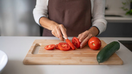 Woman's hands in apron slicing a fresh tomato on wooden cutting board in kitchenの写真素材