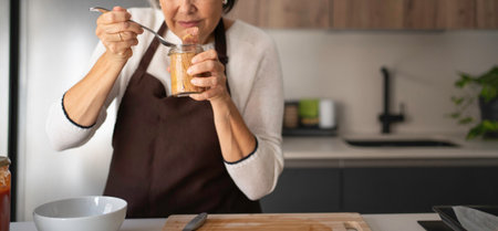 Senior woman wearing apron standing in modern kitchen, tasting directly from a jarの写真素材