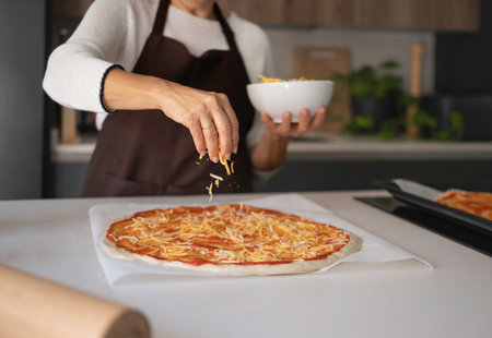 Woman's hands sprinkling shredded cheese over raw pizza dough with tomato sauce at home kitchenの写真素材