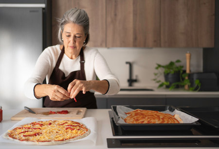 Senior woman topping homemade pizza with ingredients on a kitchen counterの写真素材