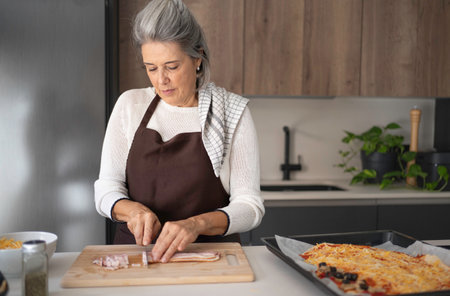Senior woman slicing bacon on a wooden board for homemade pizza in a modern kitchenの写真素材