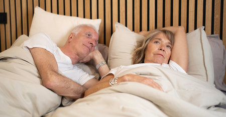 Older couple lying in bed feeling worried and thoughtful about their issuesの写真素材