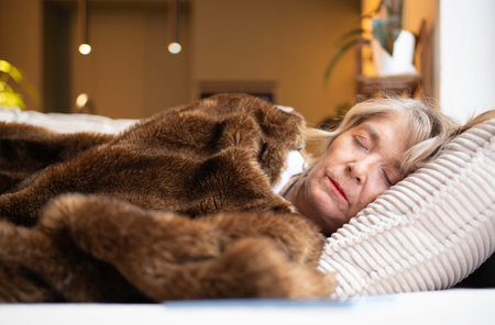 Senior woman resting peacefully on a pillow in bed under a soft brown fur blanketの写真素材
