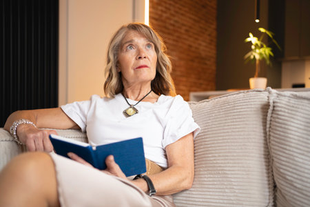 Senior woman relaxing on a sofa at home, looking up thoughtfully while holding a bookの写真素材