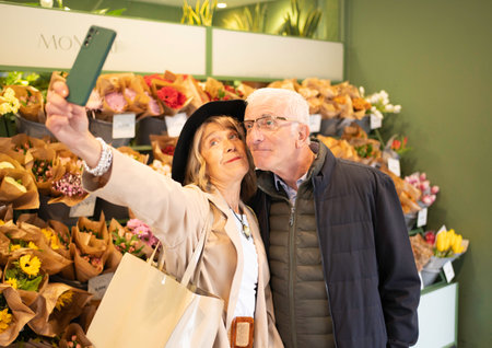 Senior couple sharing happy romantic moment taking a selfie in a flower shopの写真素材