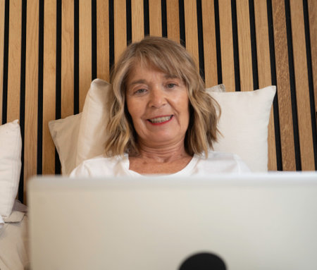 Senior woman relaxing in bed feeling happy using laptop for online communicationの写真素材