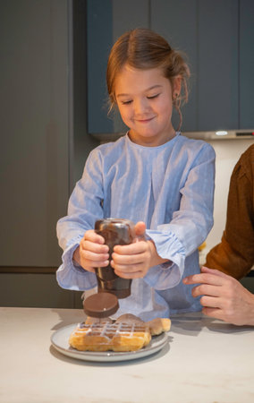 Smiling young girl happily pouring chocolate syrup on a waffle during breakfast in a kitchenの写真素材