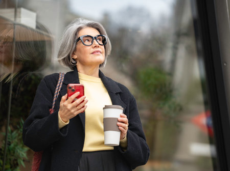 Mature woman standing contemplating with smartphone and reusable coffee cup in urban setting with...