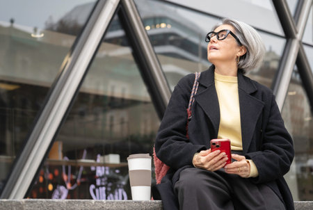 Mature woman looking up, holding smartphone and coffee cup in a modern city setting with copy space