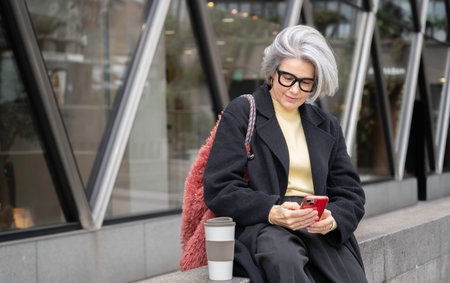 Senior woman sitting outdoors while engaging with her mobile phone and drinking coffee