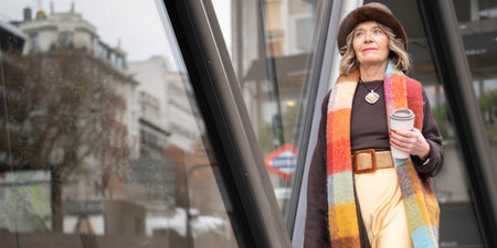 Senior woman walking in urban setting, wearing a colorful scarf and hat, holding a coffee cup