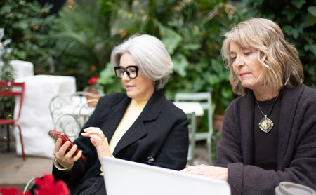 Two senior business women collaborating using a smartphone and laptop at an outdoor cafeの写真素材