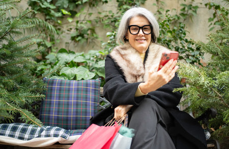 Senior woman sitting on a bench, smiling, holding shopping bags and using smartphoneの写真素材