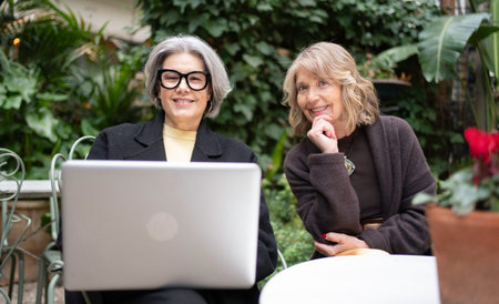 Two smiling senior women enjoying technology together on a green outdoor patioの写真素材
