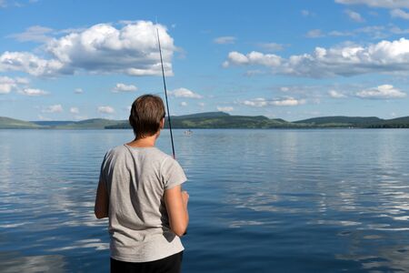 A woman fisherman stands with a fishing rod and catches fish against a blue lake and sky with clouds. Bright shot.の写真素材