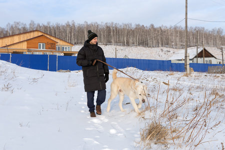 A young man walks with a big dog on a winter street in the background of village apartment...