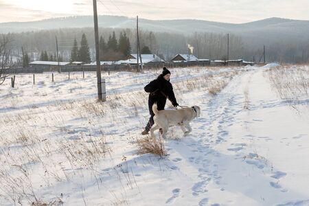 A young man runs with a big dog over a snowy field against the background of wooded mountains and village houses.の写真素材