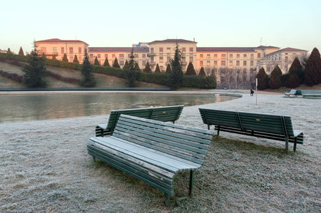 Benches stand in the Park of the Portello district on the grass in the frost on the background of a nursing home - the Palazzolo-Don Gnocchi Institute in Milan, Italy on a winter cloudy morning.のeditorial素材