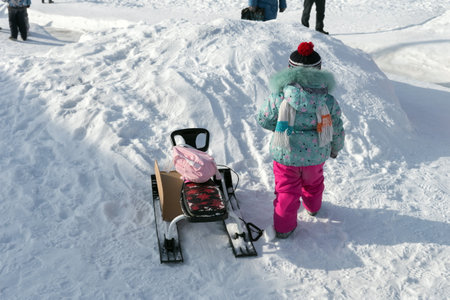 Belogorsk, Kemerovo region, RF- February 18, 2018: A child dressed in a ski suit climbs a snow slide with a snow scooter on a sunny winter day.のeditorial素材